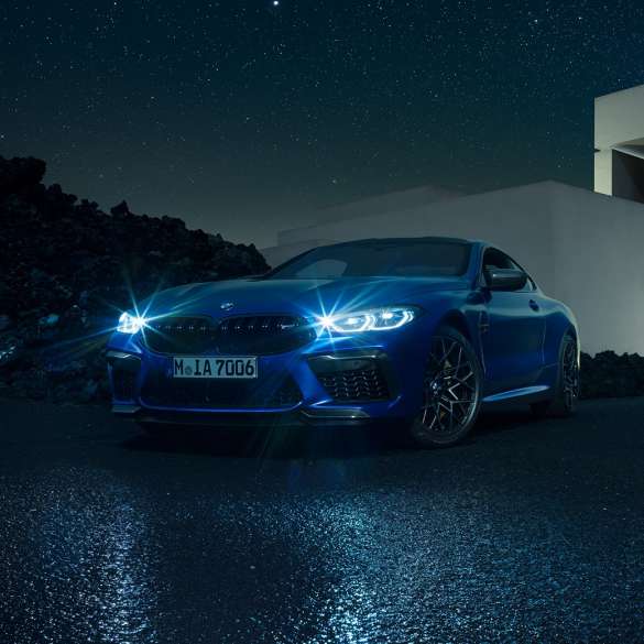 BMW M8 Competition Coupé, three-quarter front view. BMW M8 Competition Coupé in Marina Bay Blue metallic with shining LED headlights, standing three-quarter front view in front of villa at night.