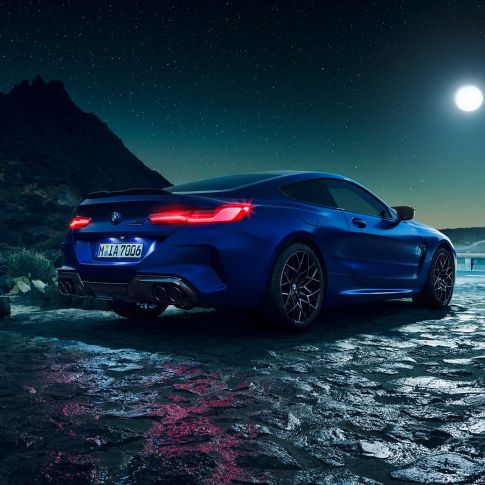 BMW M8 Competition Coupé, three-quarter rear view. BMW M8 Competition Coupé in Marina Bay Blue metallic with illuminated LED rear lights, standing three-quarter rear view with full moon.