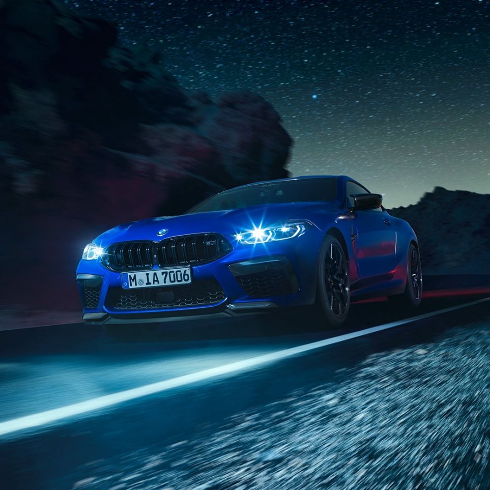Driving BMW M8 Competition Coupé, three-quarter front view. BMW M8 Competition Coupé in Marina Bay Blue metallic with illuminated LED headlights, three-quarter front view, driving at night.