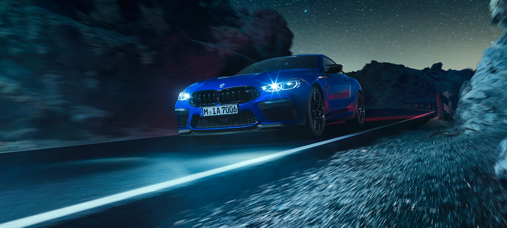 BMW M8 Competition Coupé, three-quarter front view. The BMW M8 Competition Coupé in Marina Bay Blue metallic, three-quarter front view, driving at night.