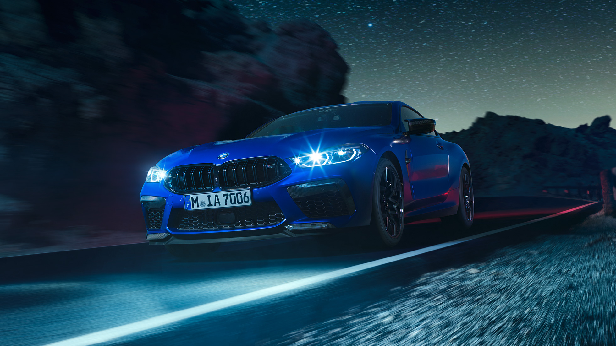 Driving BMW M8 Competition Coupé, three-quarter front view. BMW M8 Competition Coupé in Marina Bay Blue metallic with illuminated LED headlights, three-quarter front view, driving at night.