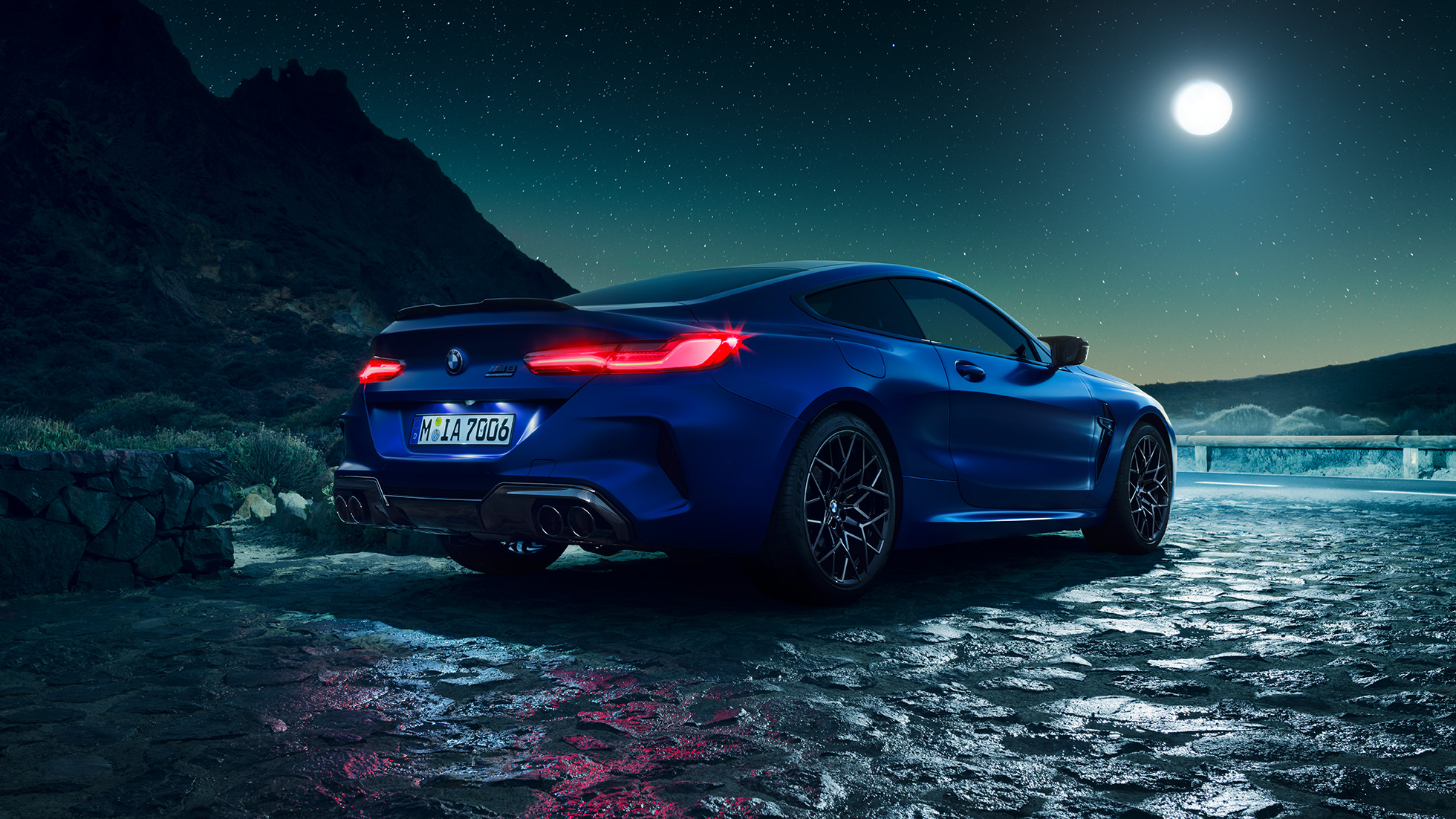 BMW M8 Competition Coupé, three-quarter rear view. BMW M8 Competition Coupé in Marina Bay Blue metallic with illuminated LED rear lights, standing three-quarter rear view with full moon.
