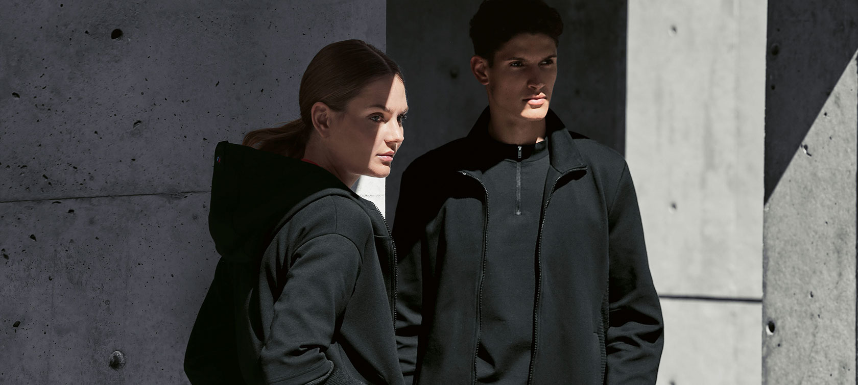 BMW M Sweat Jacket for Ladies and BMW M Sweat Jacket for Men. A couple ist standing in the shadow. A woman is presenting the BMW M Sweat Jacket for ladies and a man is wearing the BMW M Sweat Jacket for men.